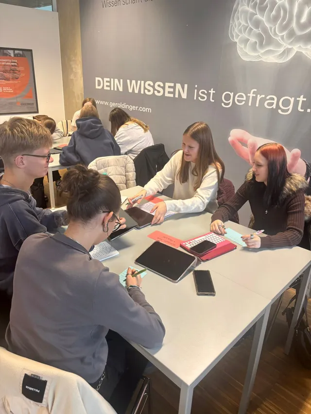 Schülerinnen und Schüler bei einer Gruppenarbeit mit Tablets, an der Wand „Dein Wissen ist gefragt.“