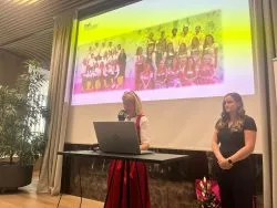 Zwei Rednerinnen am Pult bei der Maturafeier, auf der Leinwand ein Klassenfoto