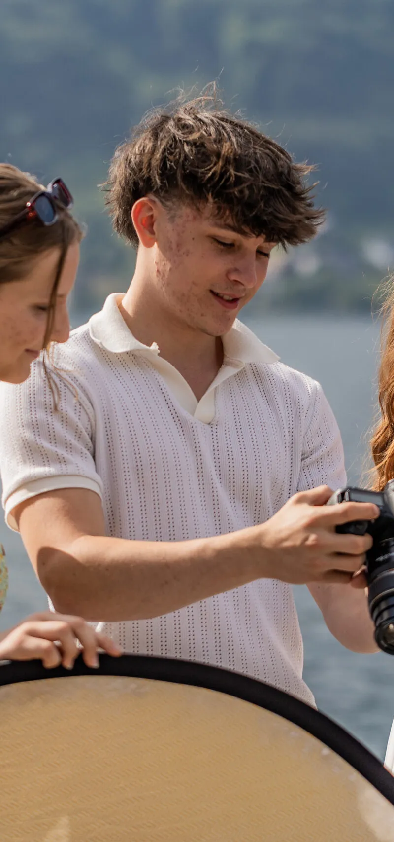 Drei Schüler:innen begutachten am Seeufer gemeinsam das Display einer Spiegelreflexkamera, eine hält einen Reflektor.
